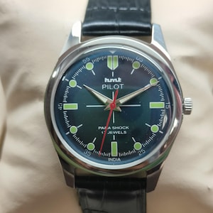 May include: A silver wristwatch with a black leather strap. The watch face is dark green with white and green markings, and the text "hmt PILOT" is visible. The watch has a red second hand and the text "PARA SHOCK 17 JEWELS INDIA".