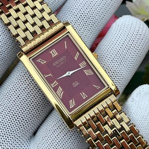 Puede incluir: Un reloj rectangular Seiko Quartz de tono dorado con una esfera color granate profundo que presenta un sutil patrón ondulado. La esfera muestra números romanos dorados, manecillas doradas y el texto Seiko Quartz SQ. Tiene una correa de metal.