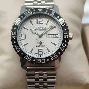 May include: A Citizen automatic wristwatch with a silver strap and a white face. The watch features a black rotating bezel with minute markers and a date display. The watch face displays the text "CITIZEN AUTOMATIC" and "21 JEWELS".