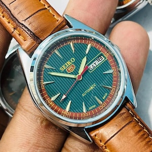 May include: A Seiko 5 automatic wristwatch with a round silver-tone case and a textured green dial featuring gold-tone indices, a day-date window, and a red second hand. The watch is attached to a brown leather strap with a crocodile-embossed pattern.