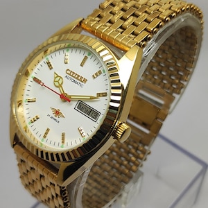 May include: A gold-tone Citizen automatic watch with a white dial, gold hands, and a red second hand. The watch features a fluted bezel, a day-date window at the three o'clock position, a 21 jewels emblem, and a gold-tone metal link bracelet.