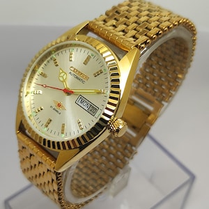 Op de afbeelding: Goudkleurig Citizen Automatic polshorloge met een champagnekleurige wijzerplaat. Het horloge heeft een goudkleurige band, een dag-datumweergave en de tekst "Citizen Automatic 21 Jewels". Het horloge wordt gepresenteerd op een transparante plastic standaard.