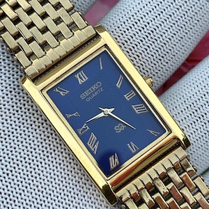 Könnte beinhalten: Eine rechteckige, goldfarbene Seiko Quartz Armbanduhr mit tiefblauem Zifferblatt, goldenen römischen Ziffern und einem strukturierten Wellenmuster. Die Uhr verfügt über ein passendes goldfarbenes Metallgliederarmband mit mehrreihigem Design.