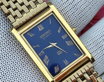 May include: A rectangular gold-tone Seiko Quartz watch with a deep blue dial featuring gold Roman numeral hour markers and a textured wave pattern. The timepiece includes a matching gold-tone metal link bracelet with a multi-row design.
