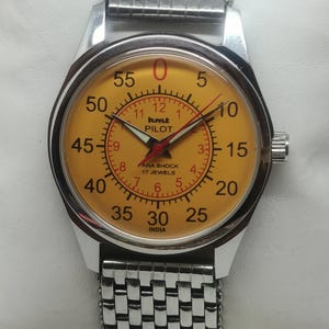Puede incluir: Un reloj HMT Pilot vintage con una correa de acero inoxidable plateado. La esfera del reloj es amarilla con números negros y agujas rojas. El texto "HMT PILOT" y "PARA SHOCK 17 JEWELS INDIA" es visible en la esfera.