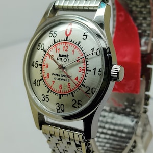 Puede incluir: Reloj de piloto plateado con esfera plateada y correa de acero inoxidable. La esfera del reloj presenta números negros y rojos, con el texto "hmt PILOT" y "PARA SHOCK 17 JEWELS". El reloj tiene una esfera redonda y una correa texturizada.