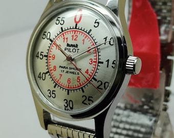 May include: Silver-toned pilot watch with a silver dial and a stainless steel band. The watch face features black and red numerals, with the text "hmt PILOT" and "PARA SHOCK 17 JEWELS". The watch has a round face and a textured band.
