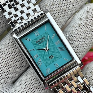 Może przedstawiać: Prostokątny zegarek na rękę Seiko Quartz z żywą turkusową tarczą z koncentrycznym wzorem fal, srebrnymi rzymskimi cyframi jako oznaczeniami godzin i srebrnymi wskazówkami, wyposażony w polerowaną metalową bransoletę z ogniwami.
