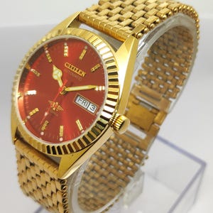May include: A gold Citizen automatic wristwatch with a red face. The watch has gold hour markers, hands, and a day-date display. The watch strap is a woven gold design. The watch is displayed on a clear plastic stand.
