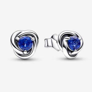 Op de afbeelding: Een paar zilveren oorknopjes met een centrale ronde blauwe edelsteen. De metalen zetting is ontworpen met een wervelend, knoopachtig patroon dat de steen omringt. De oorbellen zijn voorzien van een sluiting met een steker.