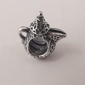 Puede incluir: Un charm de plata detallado con forma de tetera. El charm presenta intrincados patrones calados en el cuerpo, la tapa y el pico. Un pequeño botón redondo se encuentra en la parte superior de la tapa. Ideal para pulseras o collares.