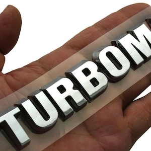May include: A chrome-finished "TURBOMAX" emblem, with a 3D effect, is displayed on a clear adhesive backing. The letters are bold and reflect light, creating a shiny appearance. The emblem is likely for automotive use.