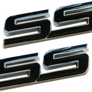 Könnte beinhalten: Zwei verchromte und schwarze SS-Embleme. Die Buchstaben sind in einer kräftigen, stilisierten Schriftart mit glänzend schwarzem Finish und Chromrand gehalten. Der Hintergrund ist weiß. Jedes Emblem ist ca. 7,6 cm lang.