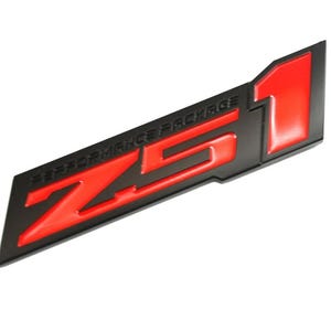 Puede incluir: Emblema negro y rojo Z51 Performance Package. El emblema presenta el texto "Z51" en rojo, con "PERFORMANCE PACKAGE" encima. El número "1" también es rojo. El emblema está sobre un fondo blanco.