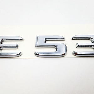 May include: Chrome-finished metal emblem spelling out "E 53". The letters and numbers are in a bold, sans-serif font, with a reflective surface. The emblem is likely for automotive use.