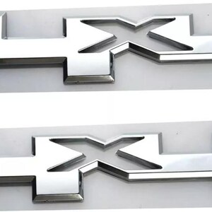 May include: Two chrome-finished 4X4 emblems. Each emblem features a stylized "4" and "X" design. The emblems are mounted on a light gray backing. The shiny, reflective surface of the emblems contrasts with the matte backing.