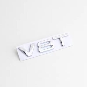 Puede incluir: Un emblema V8T cromado sobre una base rectangular blanca. Las letras son en negrita, sin serifa, con una superficie reflectante. El emblema está diseñado para aplicaciones automotrices.