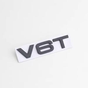 May include: A dark grey V6T emblem on a white rectangular backing. The letters are in a bold, sans-serif font, and the emblem is set against a plain white background. The emblem is likely for a vehicle.