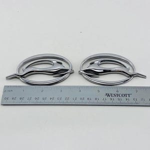 Könnte beinhalten: Zwei verchromte Impala-Emblem-Abzeichen. Jedes Abzeichen zeigt eine stilisierte springende Gazelle in einem ovalen Rahmen. Ein Lineal zeigt, dass die Abzeichen etwa 7,6 cm lang sind.