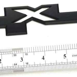 Puede incluir: Un emblema negro 4x4, de aproximadamente 7,6 cm de largo, con una regla en primer plano. El emblema es de color negro mate sólido. La regla está marcada en pulgadas y tiene la palabra "INCH" impresa.