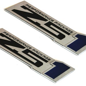 Puede incluir: Dos insignias metálicas rectangulares con el texto "NS PERFORMANCE PACKAGE" en negro. Las insignias tienen un acabado plateado con un acento azul oscuro en el extremo. Las insignias están en ángulo.