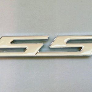 May include: A chrome-trimmed, cream-colored SS emblem. The emblem features a stylized design with two interlocking "S" shapes. The letters are filled with a glossy, off-white enamel. The emblem is likely for a vehicle.