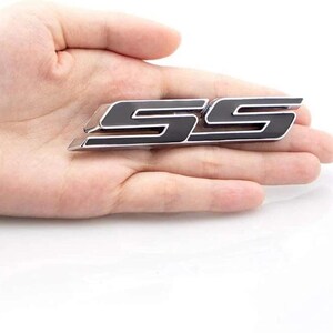 May include: A black and silver SS emblem, possibly for a car. The emblem is held in a hand against a white backdrop. The letters are stylized and have a glossy finish.