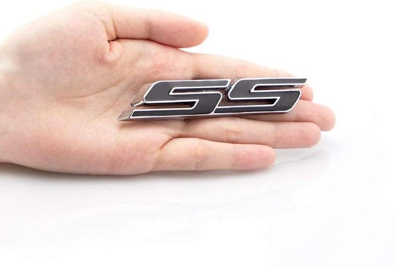 May include: A black and silver SS emblem, possibly for a car. The emblem is held in a hand against a white backdrop. The letters are stylized and have a glossy finish.