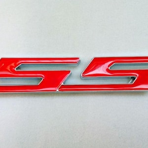 Puede incluir: Un emblema "SS" rojo brillante con un contorno plateado. El emblema presenta un diseño estilizado de las letras "S" apiladas. Las letras tienen una forma curva y angular, sobre un fondo gris claro.