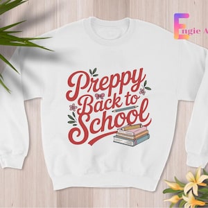 May include: White crew neck sweatshirt featuring the text "Preppy Back to School" in red script. The design includes a pencil, books, and floral accents. A casual top for the new school year.