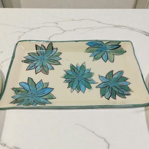 May include: Rectangular ceramic serving tray with a cream-colored base and a green border. The tray is decorated with five turquoise and blue floral designs. A decorative piece for serving food or displaying items.