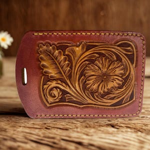 Può includere: Un'etichetta per bagagli in pelle bordeaux con un design floreale e a foglie. Il motivo in rilievo presenta un grande fiore e fogliame nei toni del marrone e del beige. L'etichetta ha un bordo cucito e una fessura per un cinturino.