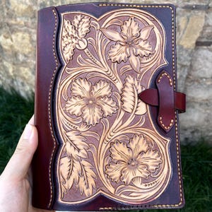 Puede incluir: Un diario de cuero artesanal con un diseño floral. La cubierta es de color burdeos oscuro con un patrón floral de color beige claro. El diario tiene un cierre de correa de cuero.