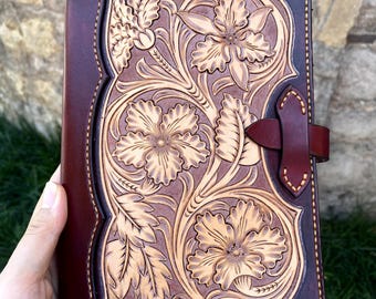 May include: A handcrafted leather journal with a floral design. The cover is a deep burgundy color with a lighter tan floral pattern. The journal has a leather strap closure and is held by a hand.