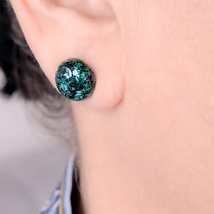 Peut inclure: Gros plan sur une boucle d'oreille ronde et verte foncée avec des accents scintillants. La boucle d'oreille est placée sur le fond d'une oreille et d'une chemise bleue et blanche à motifs. L'URL de la boutique RouteVelvet est visible en bas.