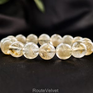 Op de afbeelding: Een armband met doorschijnende, ronde, geel getinte kralen. De kralen zijn aan elkaar geregen en vormen een cirkelvorm. De achtergrond is een donker, wazig oppervlak. De tekst "RouteVelvet" en een websiteadres zijn zichtbaar.