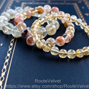 Op de afbeelding: Drie armbanden met edelsteen kralen in verschillende kleuren, waaronder heldere, gele en roodbruine stenen. De armbanden liggen op een donkerblauwe ondergrond met gouden decoratieve randen. De tekst "RouteVelvet" en een websiteadres zijn zichtbaar.