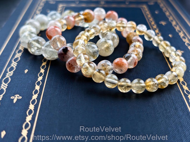 Op de afbeelding: Drie armbanden met edelsteen kralen in verschillende kleuren, waaronder heldere, gele en roodbruine stenen. De armbanden liggen op een donkerblauwe ondergrond met gouden decoratieve randen. De tekst "RouteVelvet" en een websiteadres zijn zichtbaar.