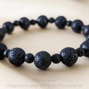 May include: A black lava stone bracelet. The bracelet features round, porous beads of varying sizes strung together. The beads have a matte finish and a dark, charcoal color. The bracelet is displayed on a light beige surface.