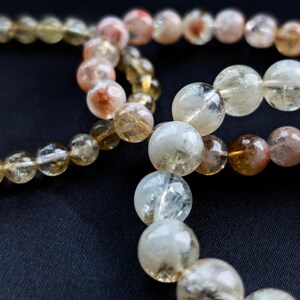 Op de afbeelding: Close-up van verschillende strengen edelsteen kralenarmbanden. De kralen zijn voornamelijk helder, met variaties van geel, oranje en bruin. De kralen zijn rond en lijken aan elkaar geregen te zijn. De achtergrond is effen zwart.