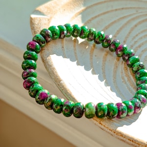 May include: A bracelet made of green and magenta gemstone beads. The beads are round and strung together. The bracelet is shown on a light-coloured, textured surface. The beads are approximately 0.8 cm in diameter.