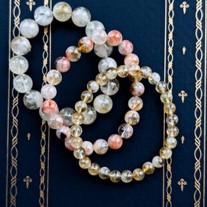Op de afbeelding: Drie armbanden met kralen van heldere, gele en rood-oranje stenen liggen op een donkerblauwe ondergrond. De armbanden zijn gemaakt van ronde, gepolijste kralen. De achtergrond is donkerblauw met gouden decoratieve randen.