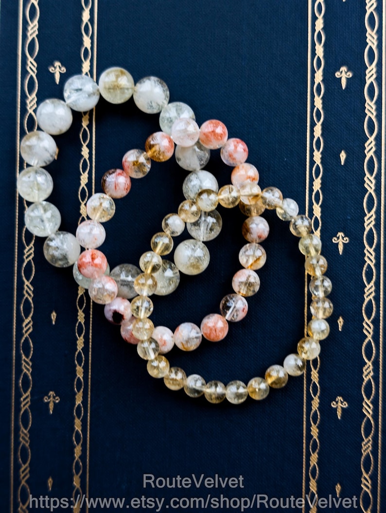 Op de afbeelding: Drie armbanden met kralen van heldere, gele en rood-oranje stenen liggen op een donkerblauwe ondergrond. De armbanden zijn gemaakt van ronde, gepolijste kralen. De achtergrond is donkerblauw met gouden decoratieve randen.