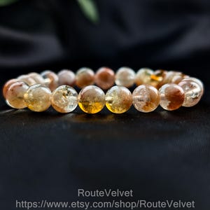 Op de afbeelding: Een armband met ronde, doorschijnende kralen in amber, bruin en helder. De kralen zijn aan elkaar geregen. De armband wordt gepresenteerd tegen een donkere achtergrond. De kralen hebben een diameter van ongeveer 1 cm.