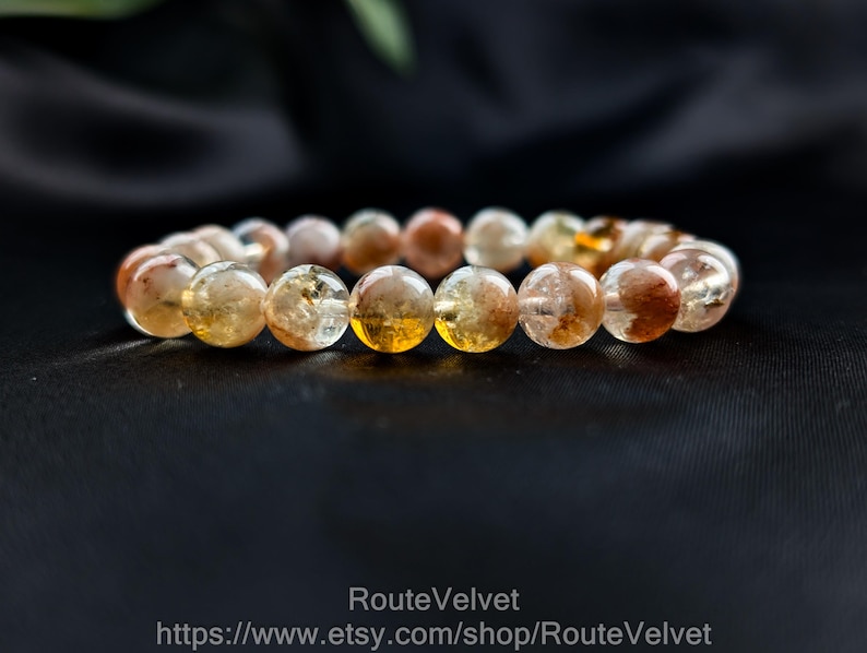 Op de afbeelding: Een armband met ronde, doorschijnende kralen in amber, bruin en helder. De kralen zijn aan elkaar geregen. De armband wordt gepresenteerd tegen een donkere achtergrond. De kralen hebben een diameter van ongeveer 1 cm.
