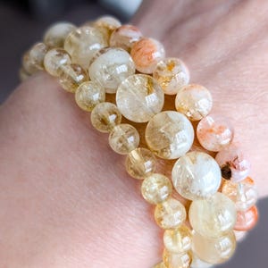 Op de afbeelding: Drie armbanden met doorschijnende, goudgele en oranje kralen van verschillende grootte. De kralen zijn aan elkaar geregen, waardoor een gelaagd effect ontstaat. De armbanden worden om een pols gedragen.