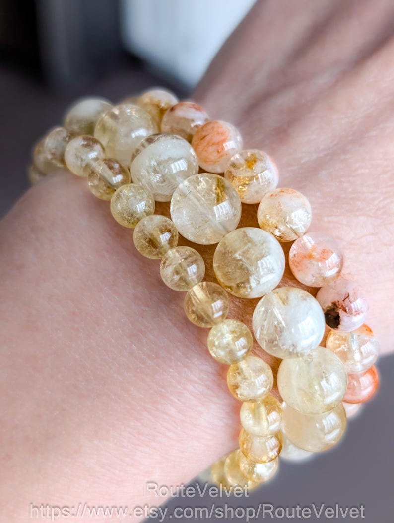 Op de afbeelding: Drie armbanden met doorschijnende, goudgele en oranje kralen van verschillende grootte. De kralen zijn aan elkaar geregen, waardoor een gelaagd effect ontstaat. De armbanden worden om een pols gedragen.