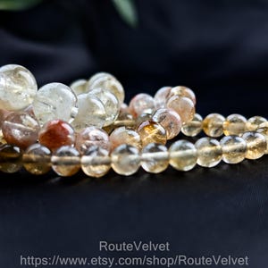 Op de afbeelding: Close-up van verschillende armbanden met kralen. De armbanden zijn gemaakt van ronde, doorschijnende, amberkleurige kralen. Sommige kralen hebben een roodbruine tint. De armbanden zijn gerangschikt op een donkere ondergrond, met een vage groene blad in de achtergrond.