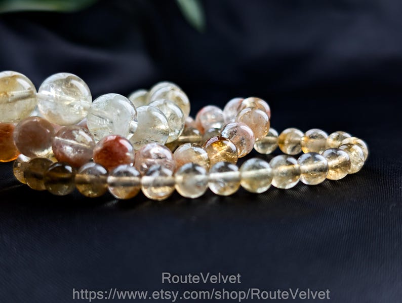 Op de afbeelding: Close-up van verschillende armbanden met kralen. De armbanden zijn gemaakt van ronde, doorschijnende, amberkleurige kralen. Sommige kralen hebben een roodbruine tint. De armbanden zijn gerangschikt op een donkere ondergrond, met een vage groene blad in de achtergrond.