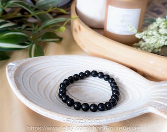 May include: A black beaded bracelet sits on a white, fish-shaped wooden tray. The bracelet is made of smooth, round beads. The background includes a wooden tray with candles and greenery.